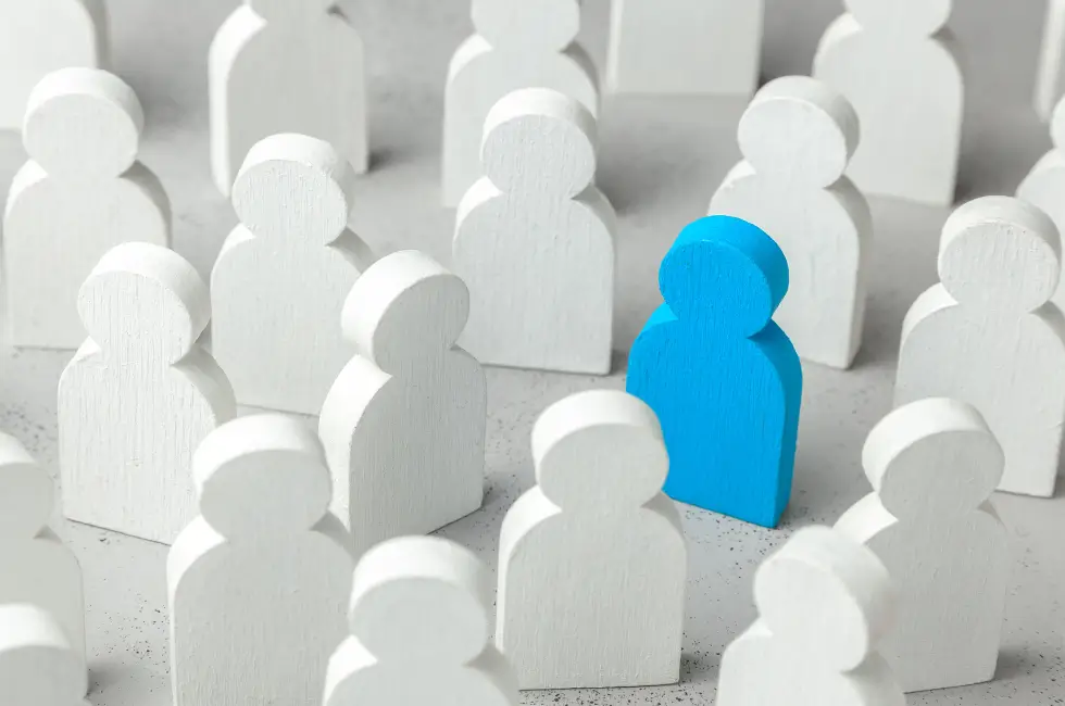 Mehrere stilisierte, menschenförmige Figuren aus Holz stehen dicht nebeneinander auf einem weißen Tisch. Die meisten Figuren sind weiß, eine einzelne Figur hebt sich durch ihre blaue Farbe deutlich von der Gruppe ab.